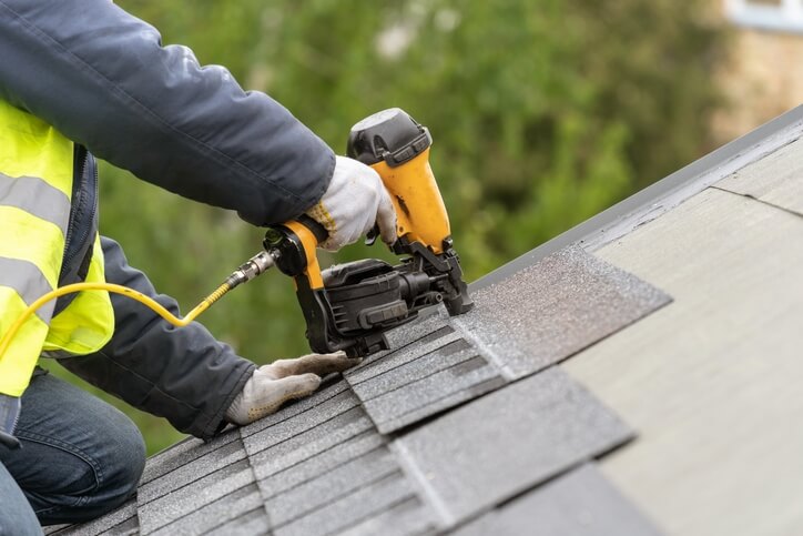 Roofing worker in a high-visibility vest using a pneumatic nail gun to fasten asphalt shingles on a sloped roof.