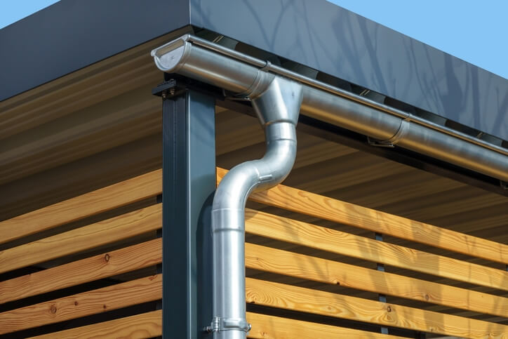 Metal downspout and gutter attached to a building corner with wooden siding and blue sky behind it.