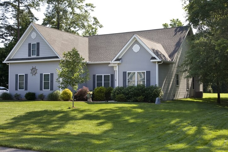 Front view of a single-story house with gray-blue siding, white trim, and a well-kept lawn with shrubs and a small tree in front.